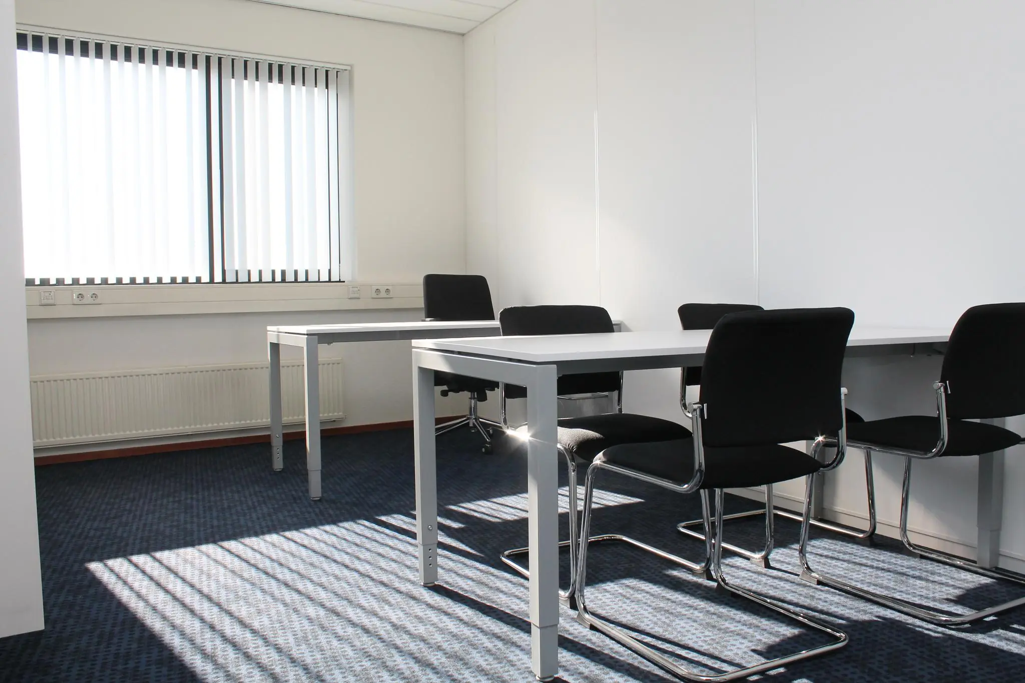 Kleine vergaderruimte aan de Laan van ’s-Gravenmade met twee tafels, vier zwarte stoelen en lichtinval door verticale jaloezieën.